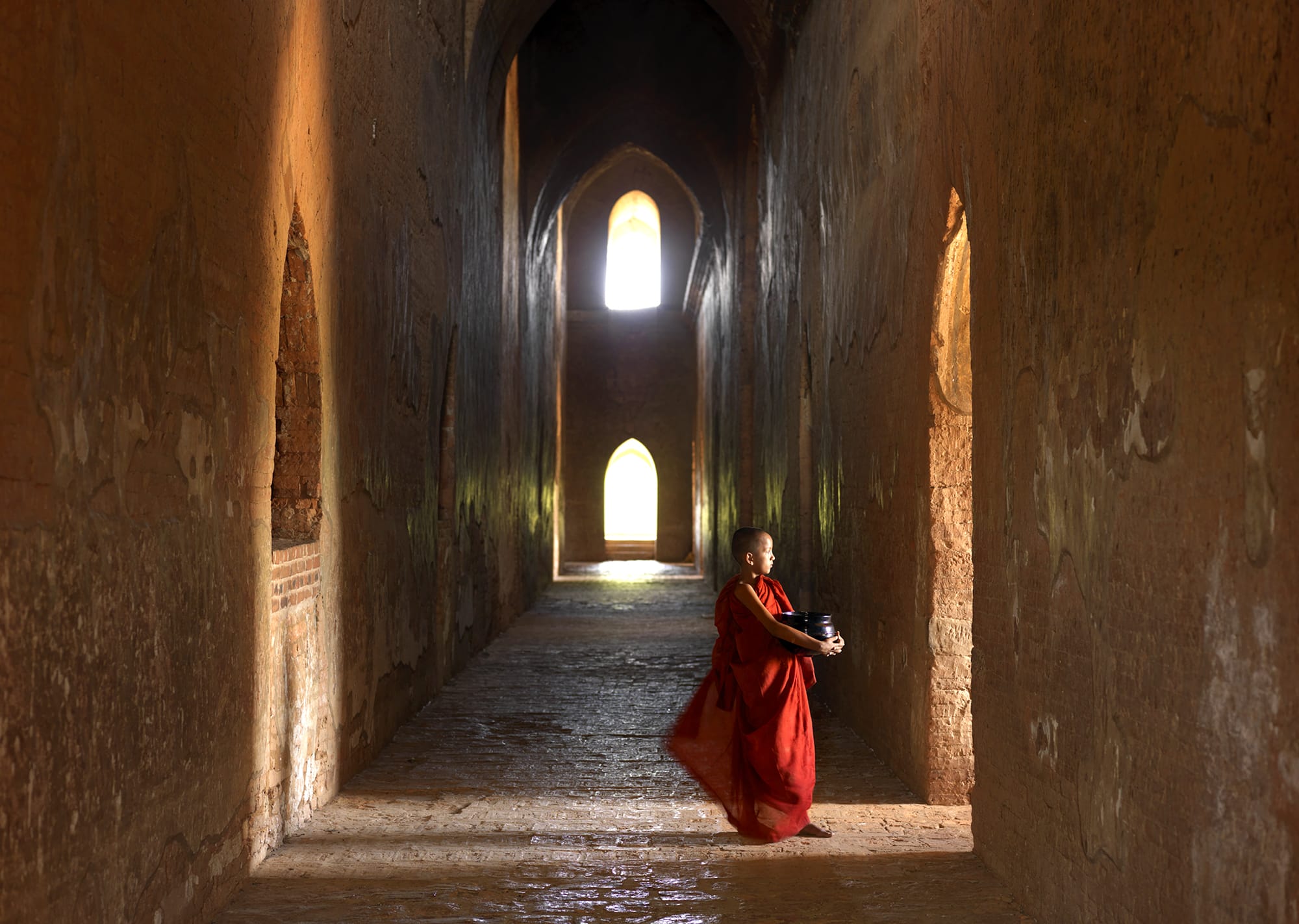 A young monk in Bagan stands in a temple corridor, his red robe billowing in the wind, illuminated by soft light and ancient stonework.