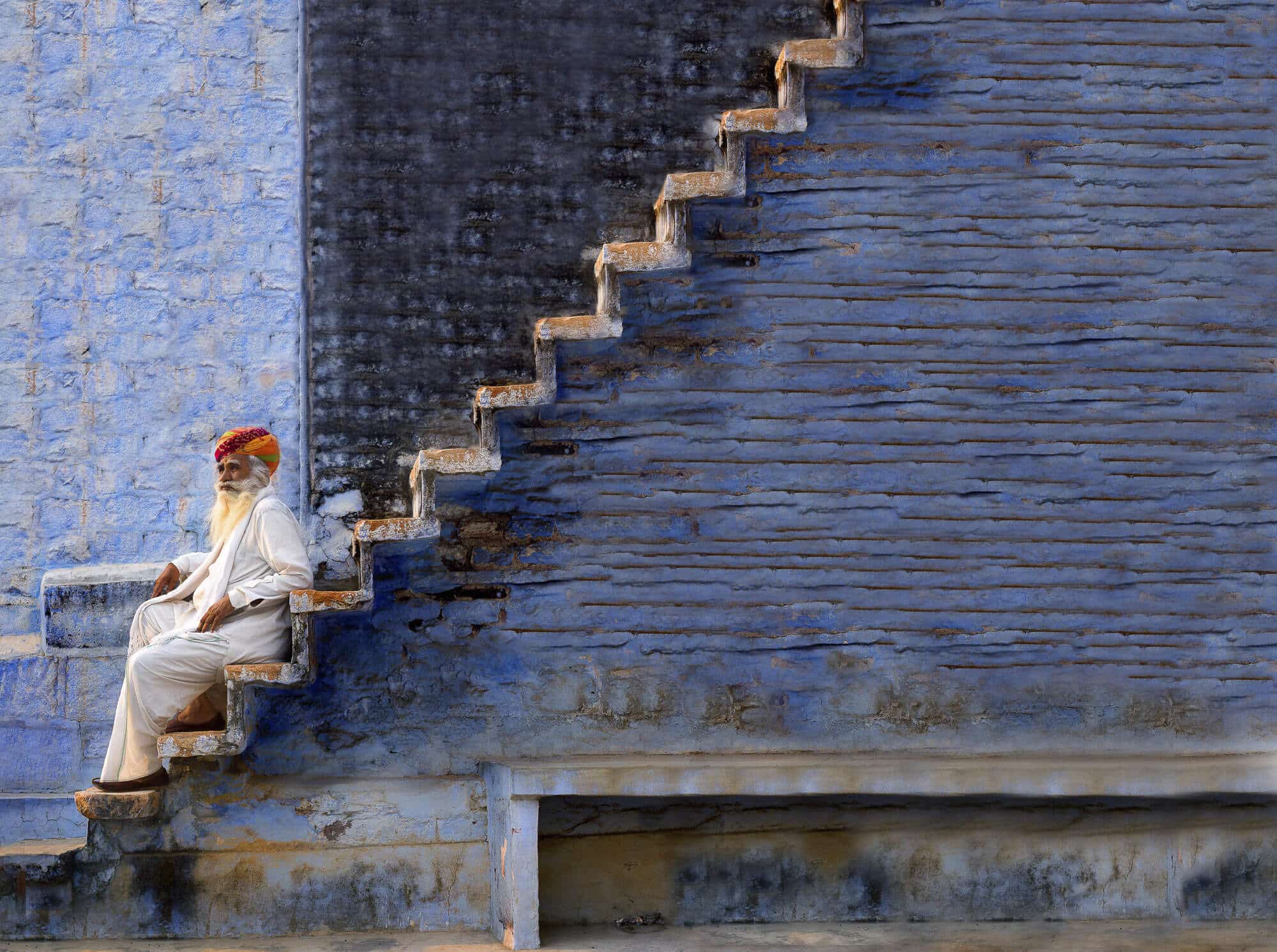 A guardian sits on ancient steps in Jodhpur, India, embodying wisdom and resilience against the backdrop of timeless blue architecture.